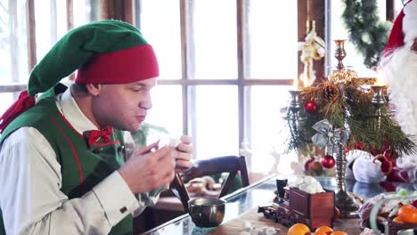 hungry male elf eating dessert at christmas. elf in green costume drink tea with pastries sitting next to santa indoors