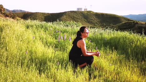 slow motion of female in black dress and sunglasses kneeling in green field and watching sunset in distance