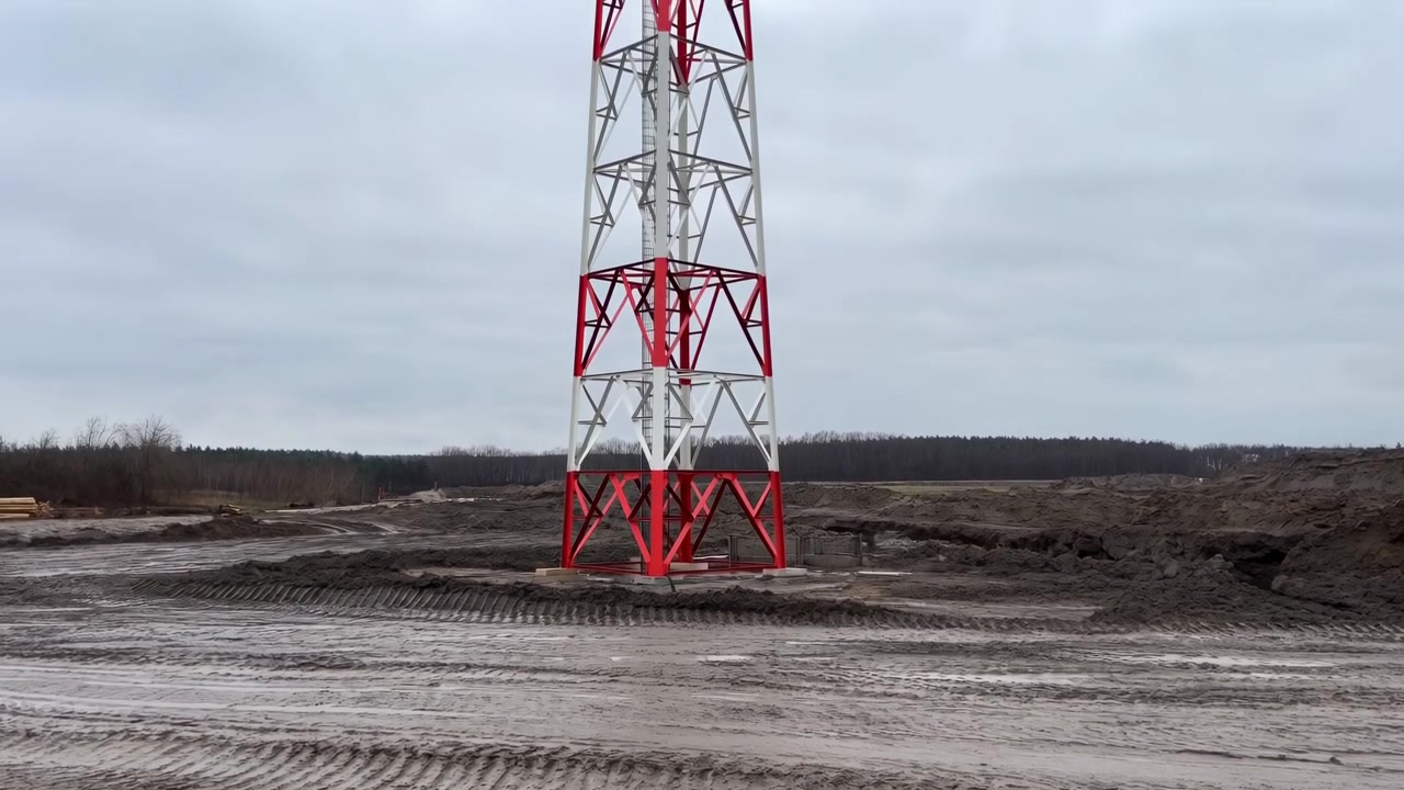 Vista completa de una torre de celosía de acero roja y blanca anclada sobre cimientos de hormigón en una obra embarrada, mostrando arriostramiento, sección de escalera, superficie metálica pintada y cielo gris.