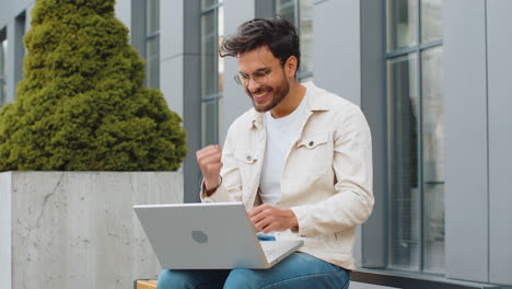happy indian man working on laptop celebrate success win money get online good news outdoors street