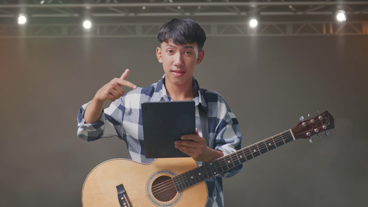 adolescente asiático con una guitarra apuntando a una tableta en la mano mientras está de pie en el escenario