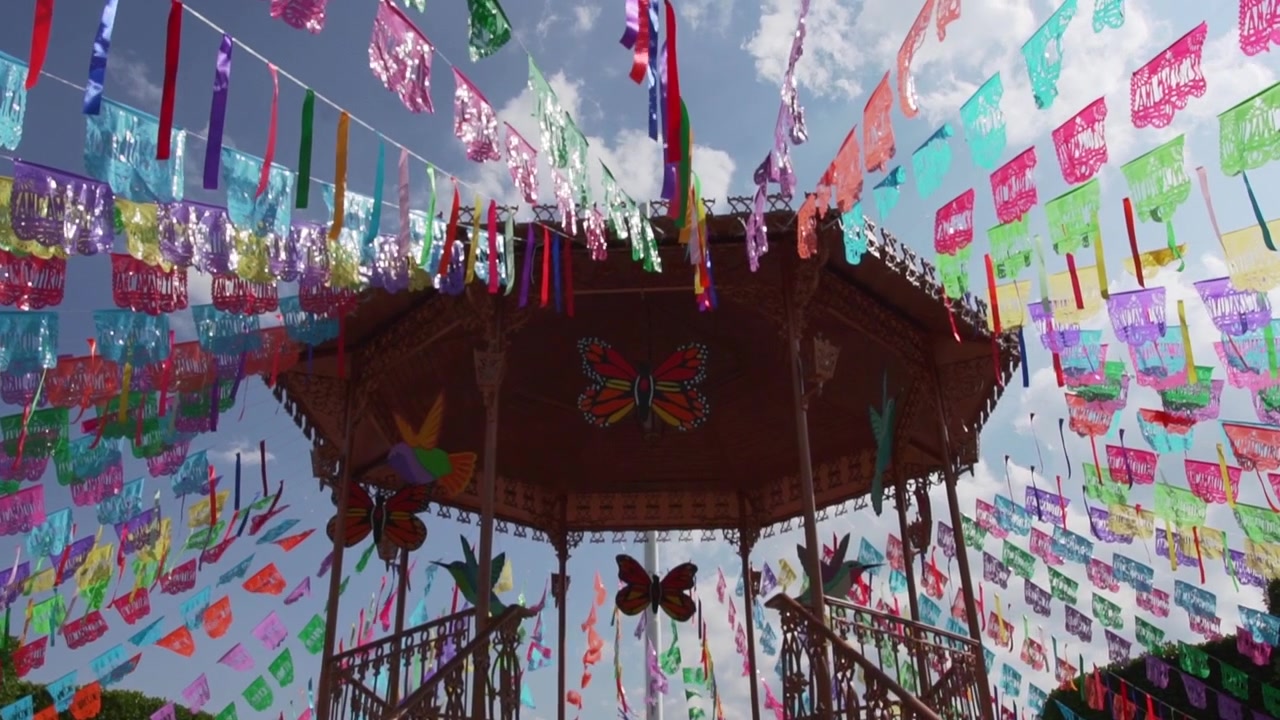 Spanduk papel picado Día de Muertos yang penuh warna di sekitar gazebo berhias, dengan ornamen kupu-kupu dan dekorasi festival Meksiko yang cerah
