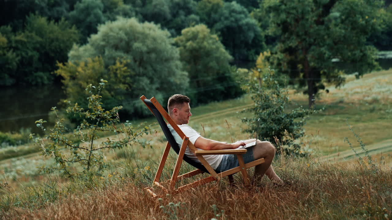 Nice place to work remote in the nature. Caucasian man works on his laptop sitting in a folding chair. Side view.