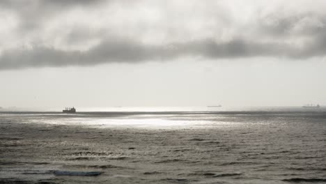 Una-Escena-Oceánica-En-Calma-Con-Nubes-Oscuras-Arriba-Y-Charcos-De-Luz,-Barcos-Que-Pasan-Lentamente-Por-El-Horizonte