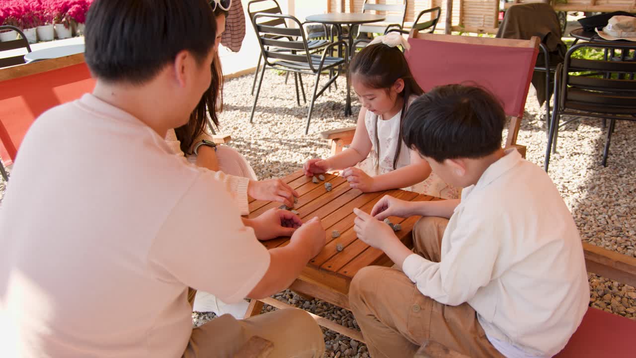 日当たりの良い屋外カフェのテーブルで、アジア系の両親と子供たちが一緒にゲームをする