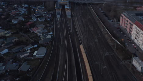 Aerial-view-of-a-passenger-train-passing-by-trees-in-Berlin,-Germany-moving-towards-station-with-people-waiting-under-a-bridge-with-cars-moving-surrounded-by-small-hut-houses-during-an-early-morning