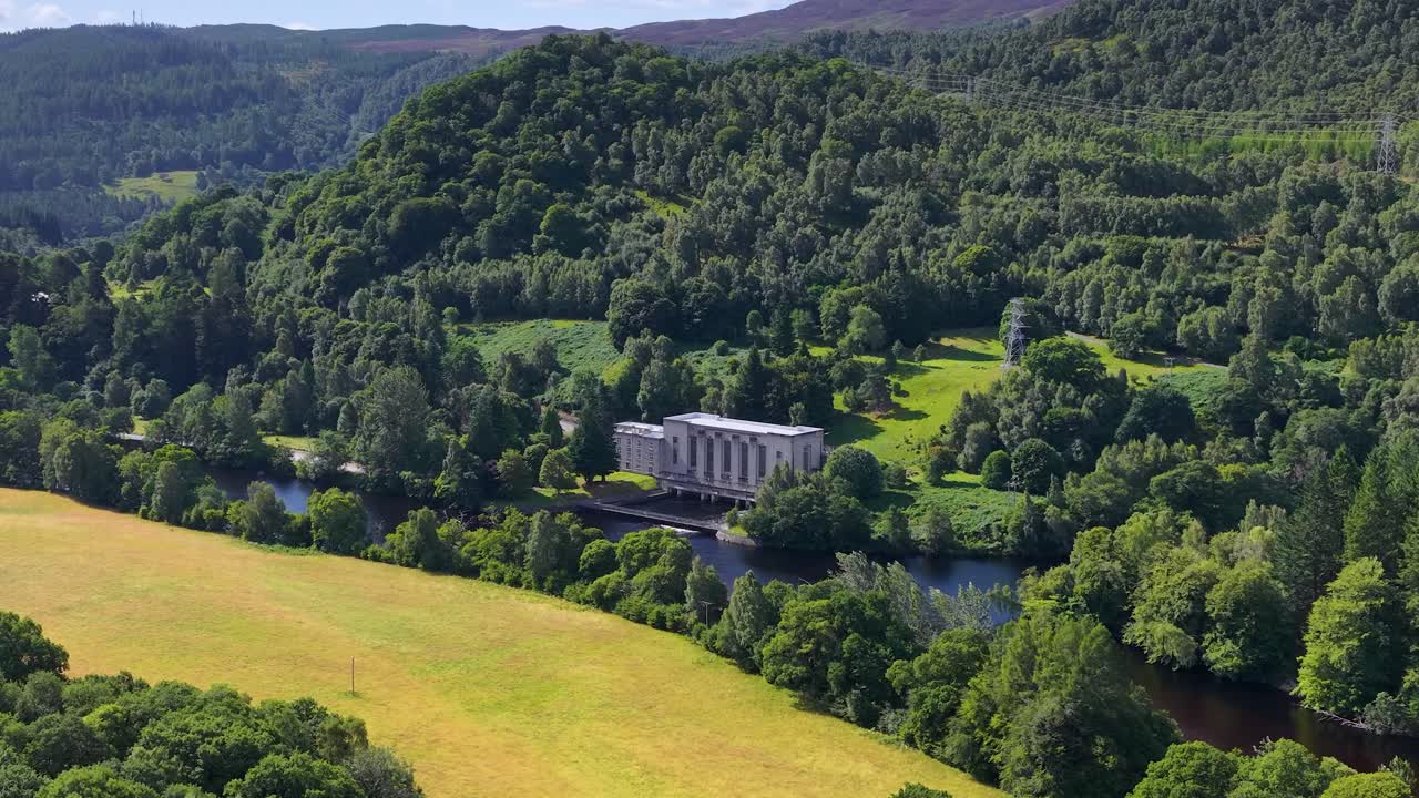 Drone glides over lush green landscape toward hydroelectric station by river, bright daylight, smooth motion