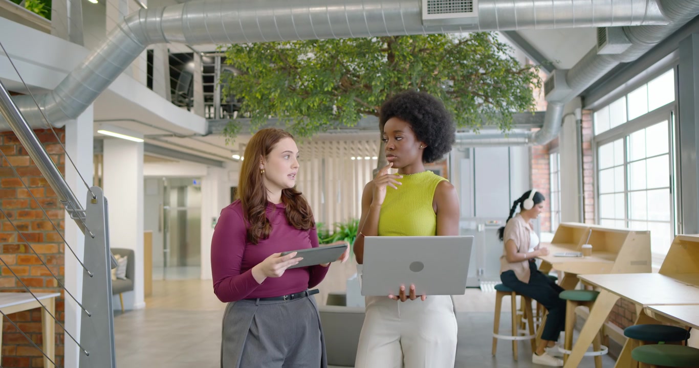 Two female coworkers in office tapping tablet gesturing and discussing with laptop finalizing files