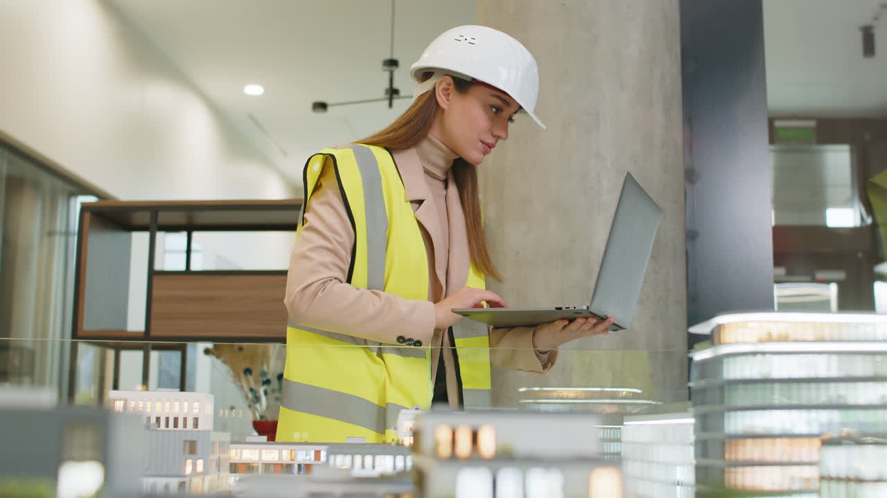 Confident architect in hardhat using laptop while analyzing office complex model at workplace