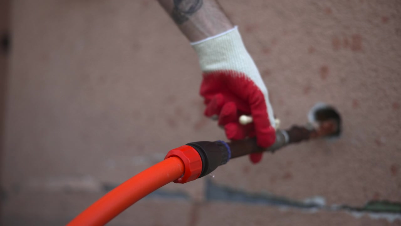 Primo piano di un operaio che indossa un guanto protettivo mentre leviga intonaco fresco su una parete non finita con una cazzuola, con un tubo arancione nelle vicinanze, in un contesto di ristrutturazione.