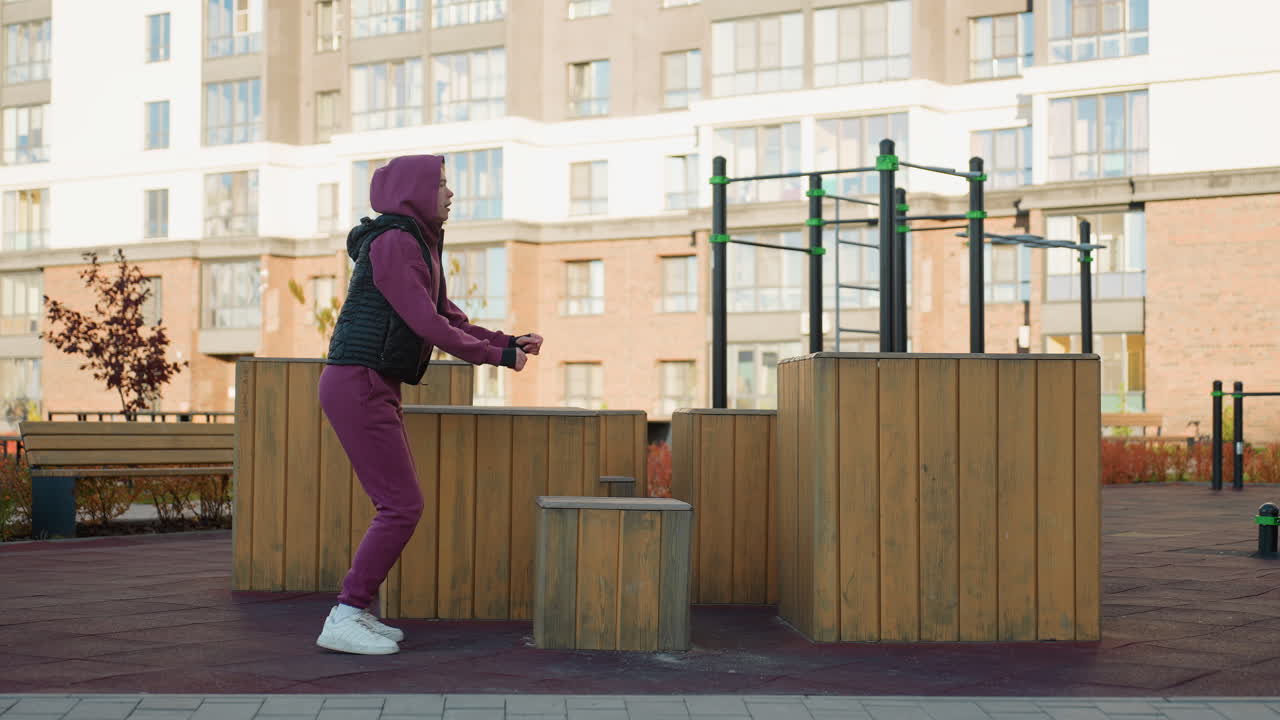 Strong willed trainee jumps onto wooden plyo box then jumps down during intense outdoor workout in urban park showcasing explosive power balance and fitness focus under clear sky city backdrop