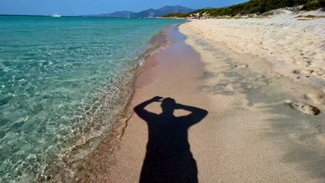 Silhouette-shadow-of-man-putting-hat-on-and-walking-on-sandy-beach-along-turquoise-sea-water-shoreline