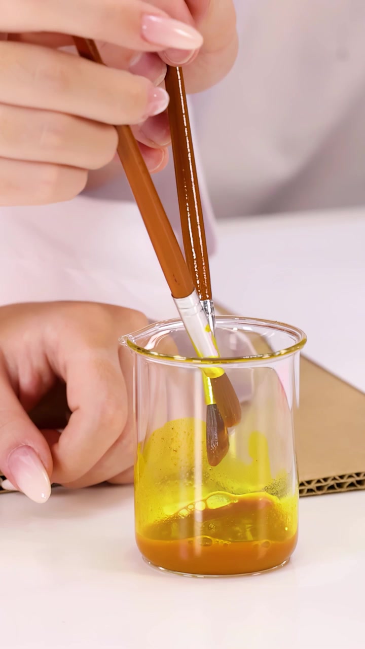 Primer plano de las manos de un estudiante mezclando líquido amarillo en un vaso de precipitados de vidrio usando pinceles para un experimento.