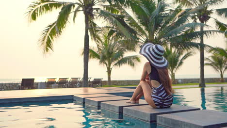 a healthy, fit, woman sitting, with her back to the camera, on the edge of a large resort swimming pool looking out at the ocean horizon