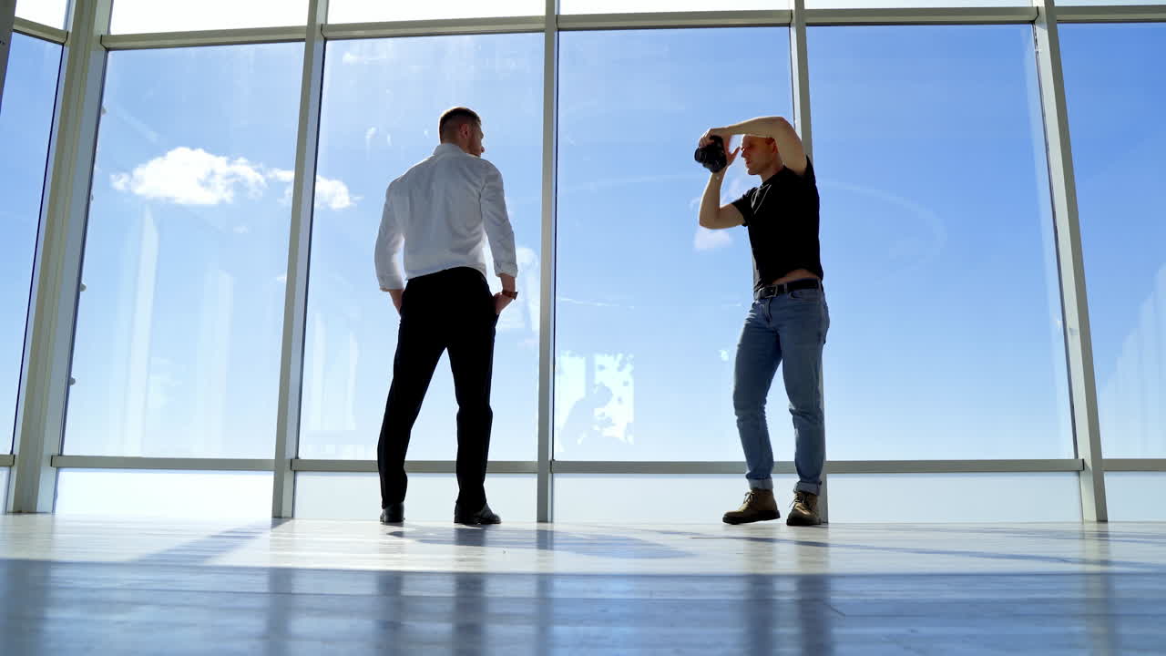 Male photographer taking photos of a businessman. Rear view of a young man in white shirt and black trousers posing on camera indoors. Panoramic windows.