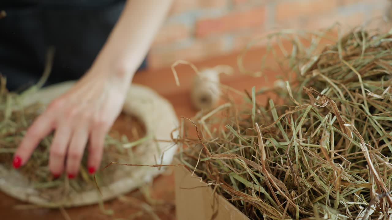 Florist hand with red manicure arranges natural hay bundle on wooden table for floral craft project, preparing organic material for decorative arrangement design in rustic workshop with creative focus