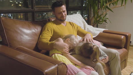 happy father sitting on sofa with his two little girls who sleeping and leaning their heads on his lap