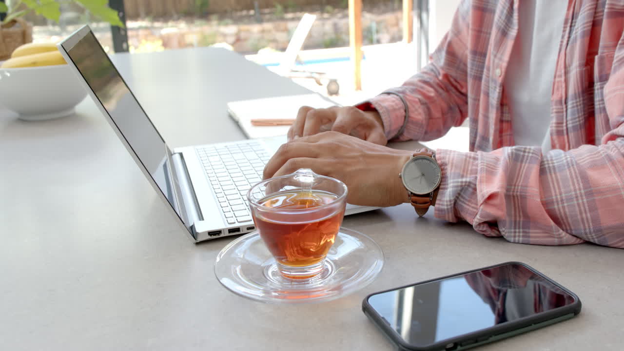 Uomo che digita sul laptop, lavorando da casa con tè e smartphone nelle vicinanze.