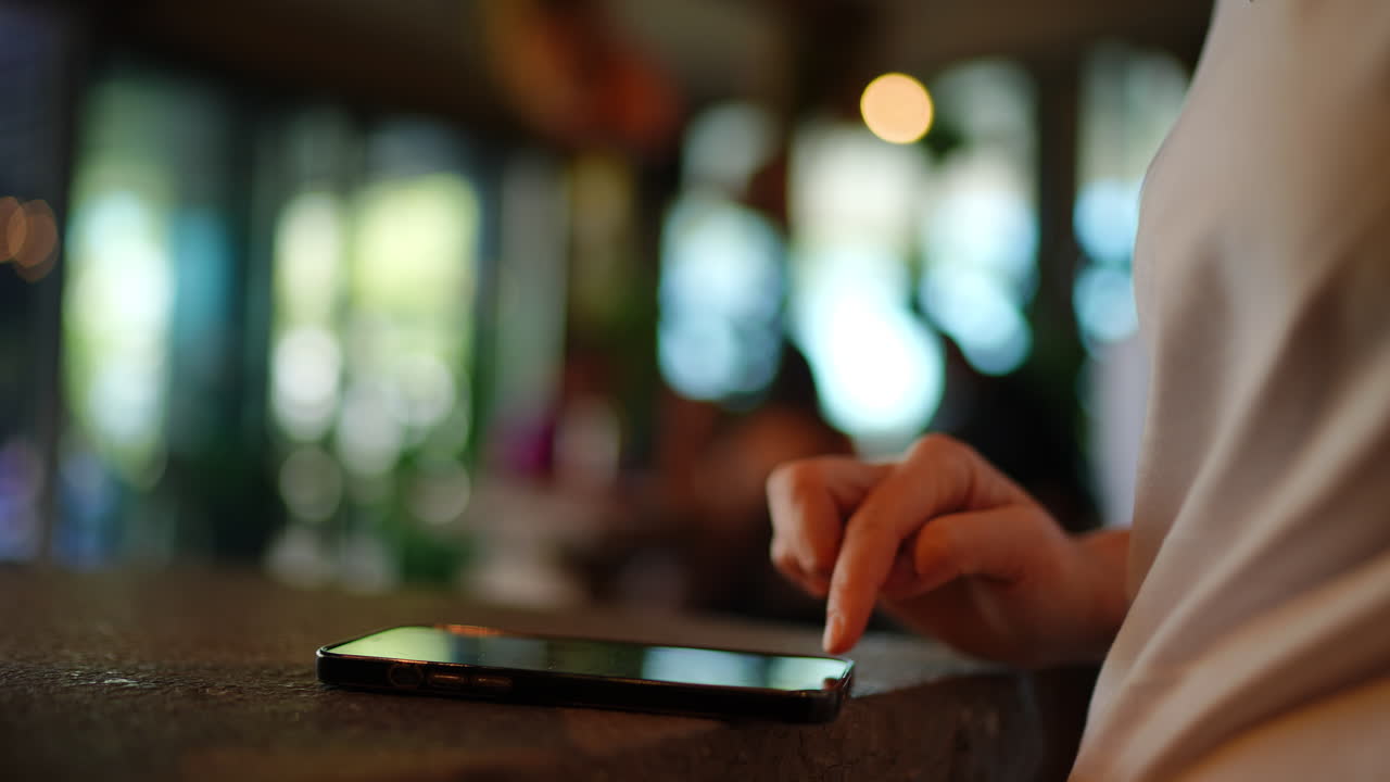 Young woman at restaurant, intently browsing smartphone screen, scrolling and tapping digital content with focused finger movements, blurred background creating soft light ambiance