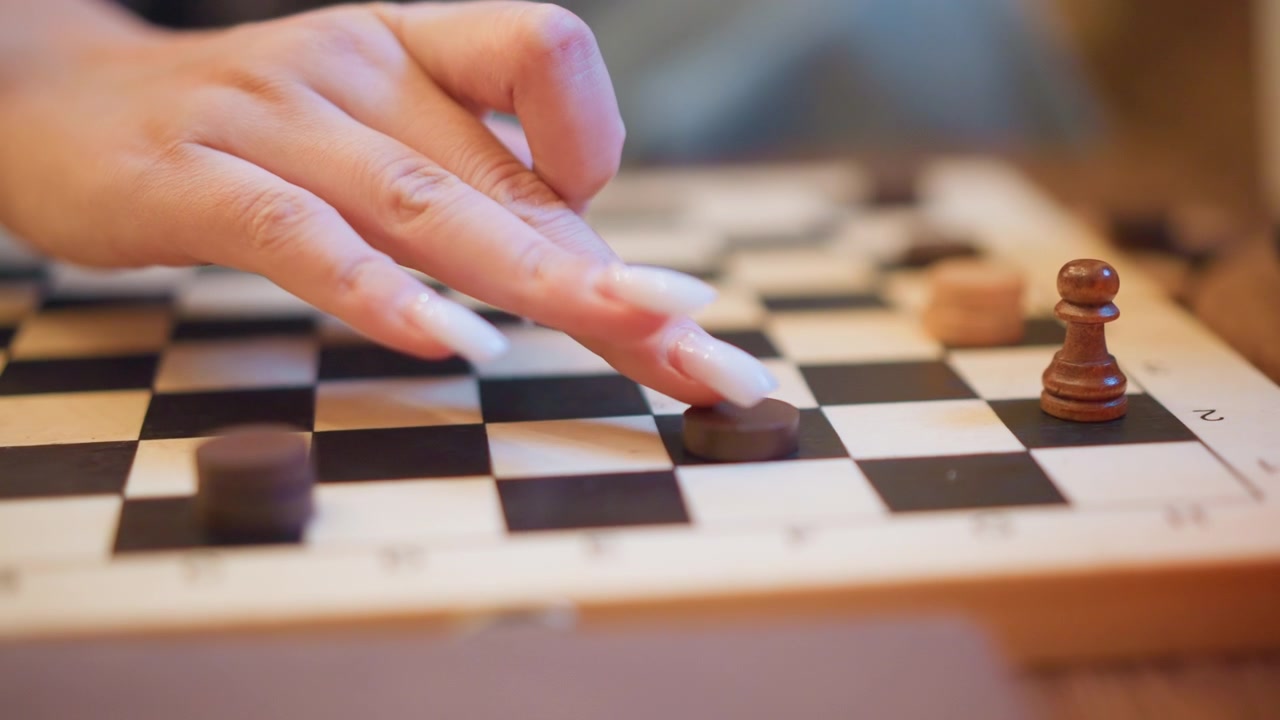 Close-up de mão com unhas longas movendo peças de damas em um tabuleiro. Um peão de madeira e casas pretas são visíveis, mostrando a progressão do jogo à medida que fichas de cores diferentes são colocadas.