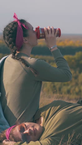 girl with drink and partner rests after day hike on autumn grass at camp in calm autumn evening closeup