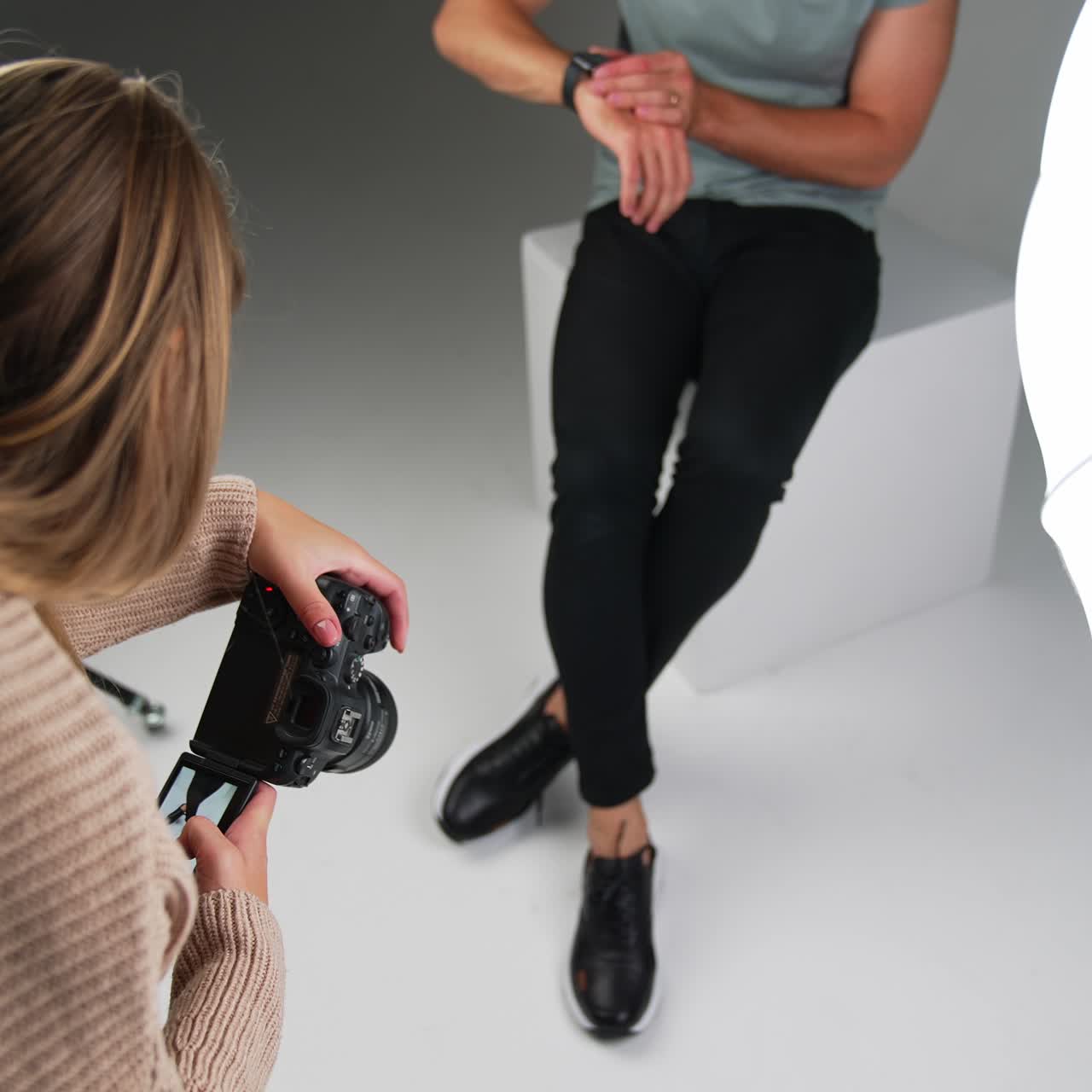 Long-haired woman photographer taking picture on a professional camera. Studio footage of a male model sitting on a white cube