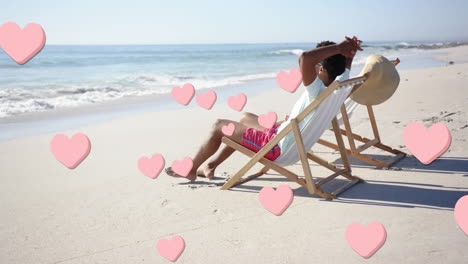 animation of pink hearts over african american man relaxing on beach