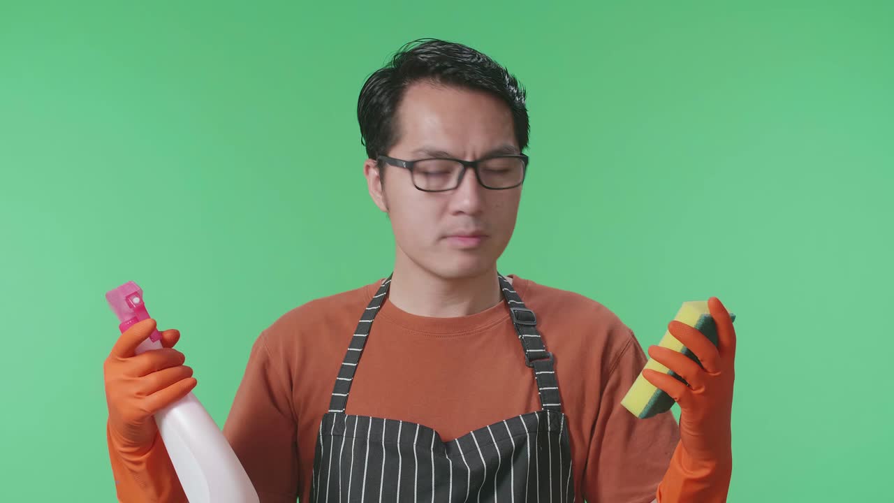 손에 스프레이와 주방 스폰지를 들고 있는 애시안 남성 집주인이 초록색 스크린 스튜디오에서 머리를 흔들고 있다.