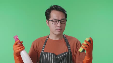primer plano de ama de casa masculina asiática con un delantal mirando spray y esponja de cocina en las manos luego sacudiendo la cabeza en el estudio de pantalla verde