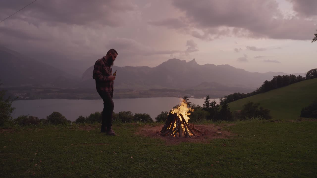 A man stands near a burning campfire on a grassy hill with mountains and a lake in the background at sunset. Concept of solitude, reflection, wilderness adventure, and peaceful living