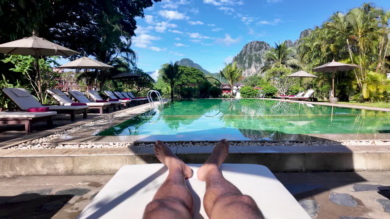 A person is relaxing on a sun lounger by the swimming pool at a boutique resort in Vang Vieng Laos The scene features mountain views and a blue sky ideal for travel and tourism related content