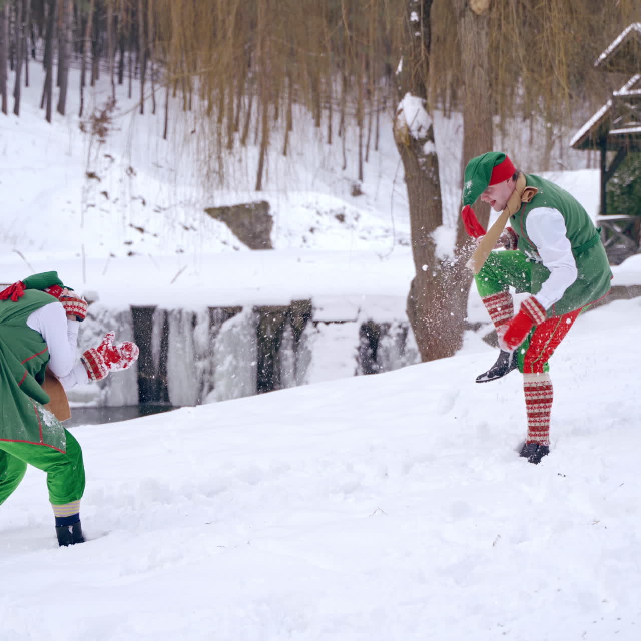 Funny elves play snowballs in nature. Snowy background and two joyful fairy characters in green costumes having time together outdoors. Christmas.