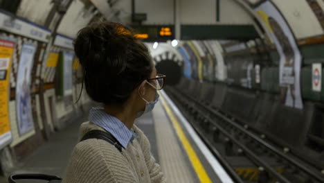 woman wearing a face mask waits in an empty tube station, london, uk