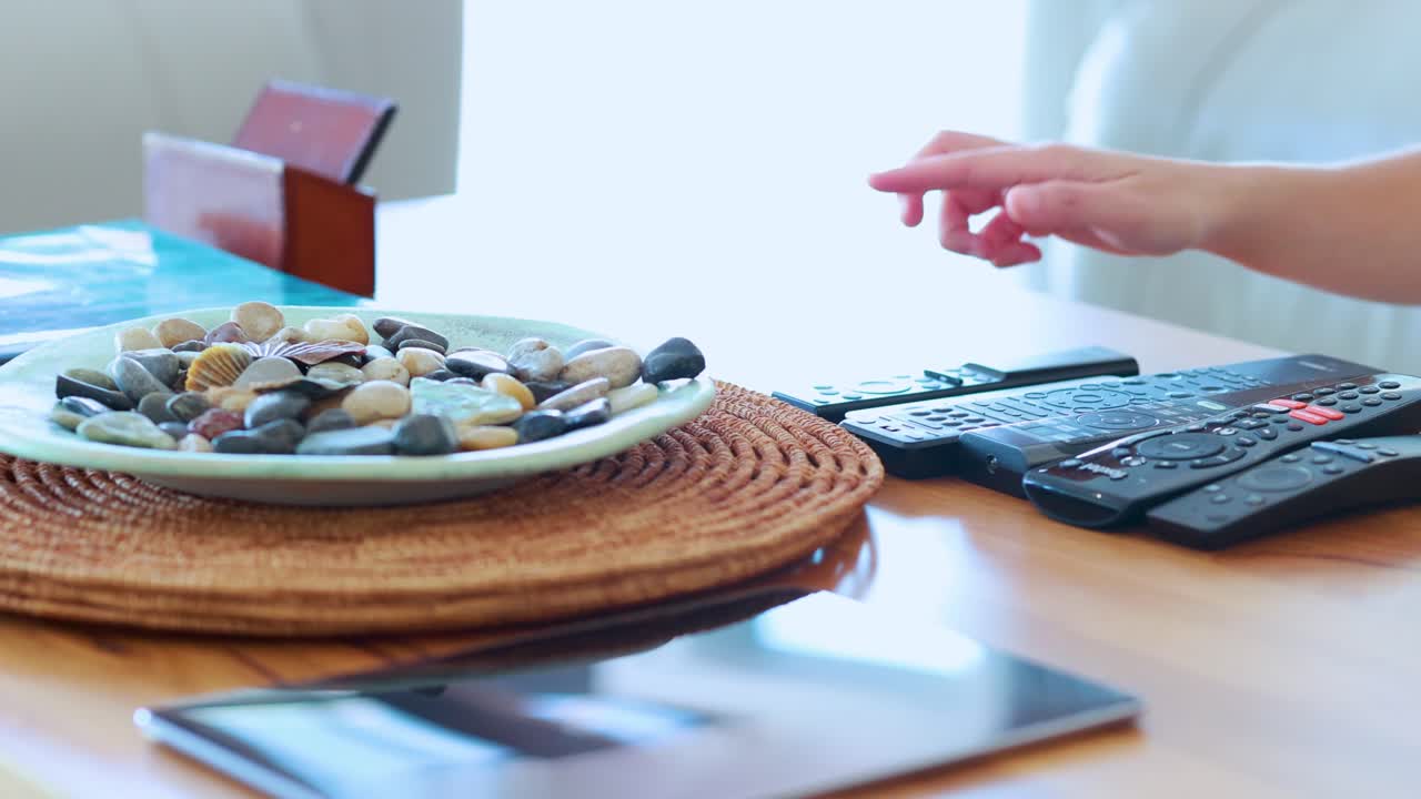 A hand reaches for and lifts a remote control from a modern coffee table with decorative stones, in soft natural daylight, static camera