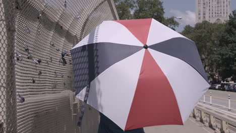 following shot of a woman walking across a bridge carrying a red white and blue umbrella on a sunny day