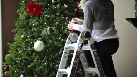 Niña-En-Una-Escalera-Decorando-El-árbol-De-Navidad-Con-Luces-Navideñas,-Bolas-Y-Flor-De-Nochebuena-Para-La-Temporada-Navideña