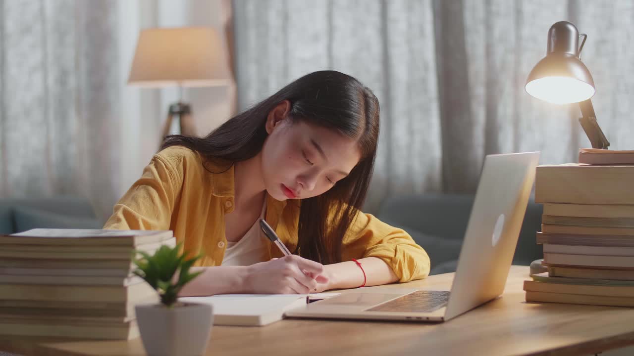 젊은 아시아 여성이 집에서 노트북으로 공부하는 동안 책에 글을 쓰고 있습니다.