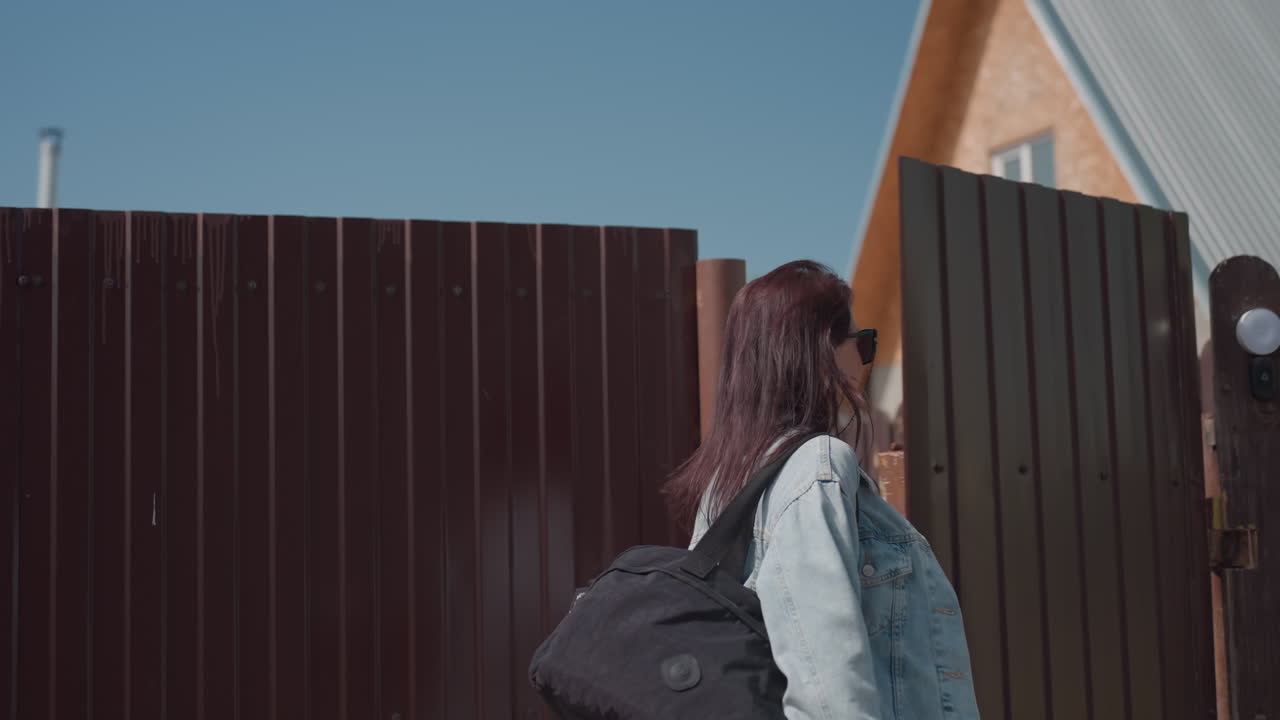 Smiling young woman in white crop top and denim walks through open wooden gate into sunny residential yard while friend follows behind, warm daylight and casual mood