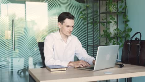 portrait of a handsome, young, stylish man in business attire, sitting at a desk with a laptop in a modern office