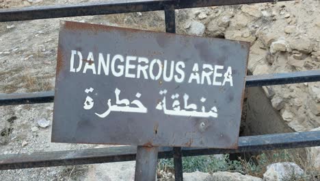 A-warning-sign-with-dangerous-area-in-English-and-Arabic-near-a-steep-drop-off-in-desert-location-of-Jordan-in-Middle-East