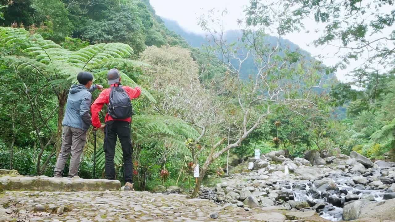 Dua pendaki berdiri memandang sungai berbatu dan pegunungan hijau subur di jalur bebatuan