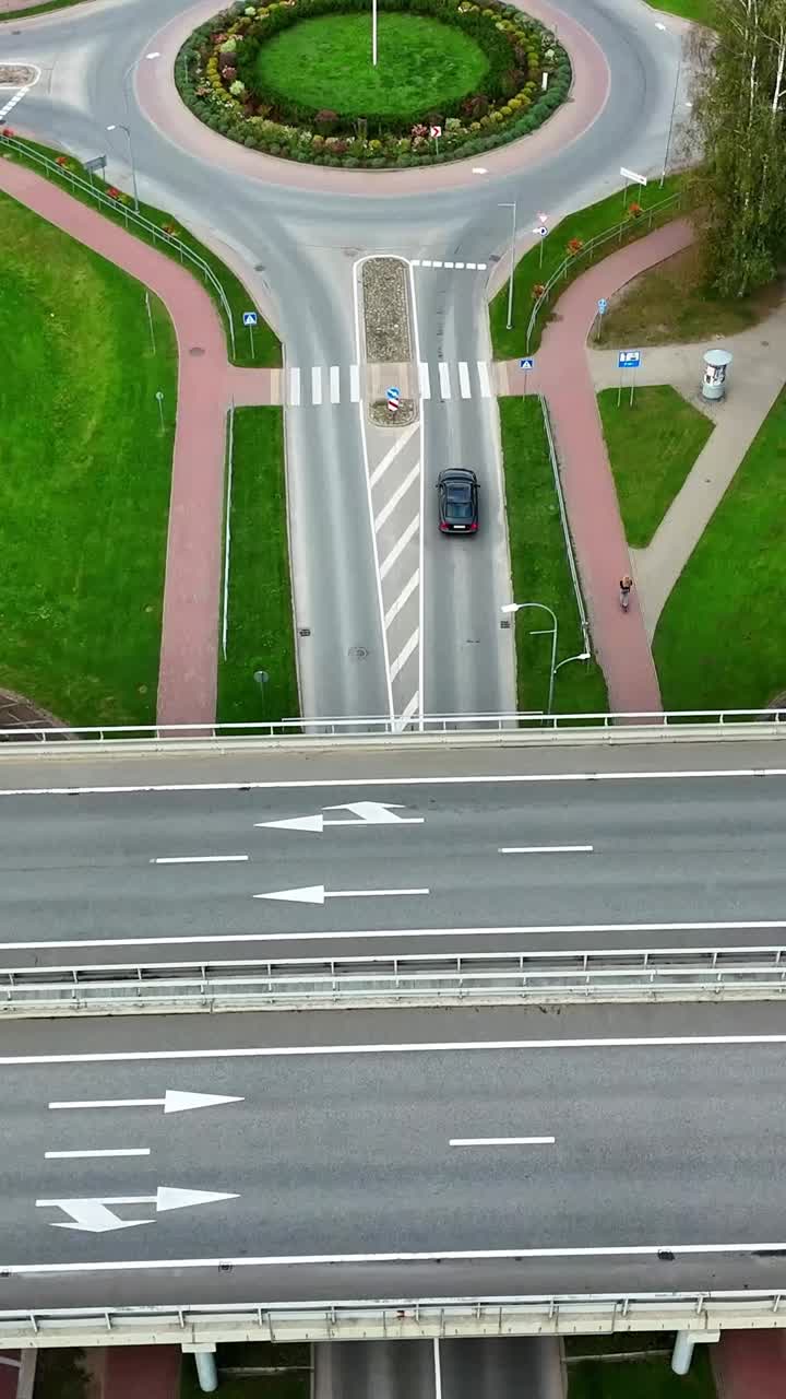 Top-down vertical drone shot of a landscaped roundabout with pedestrian crosswalks and adjacent parking lots in Ikskile, Latvia, highlighting urban planning and traffic flow