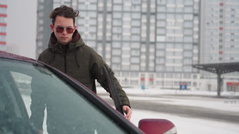 young man in winter jacket opens car door and enters vehicle amidst snowfall in snow covered parking area with blurred residential buildings in background
