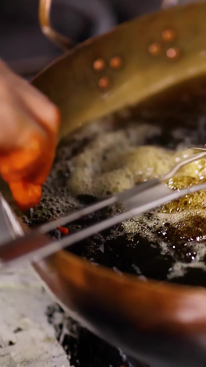 Close-up of hands dropping batter into hot oil inside a brass wok for deep frying.
