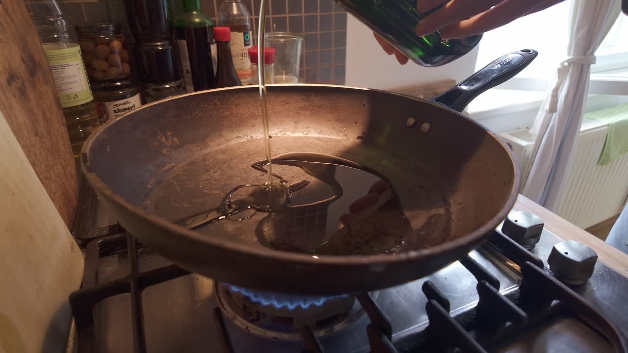 Domestic chef pouring olive oil into hot frying pan on kitchen gas hob