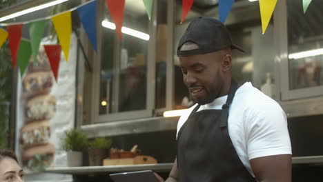 Male-Waiter-With-Tablet-Taking-Note-Of-A-Happy-Young-Couple's-Order-At-Truck-Bar