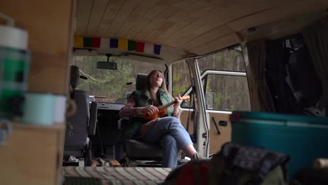 a young girl with red hair plays the ukulele inside a caravan in the countryside. travelling in.