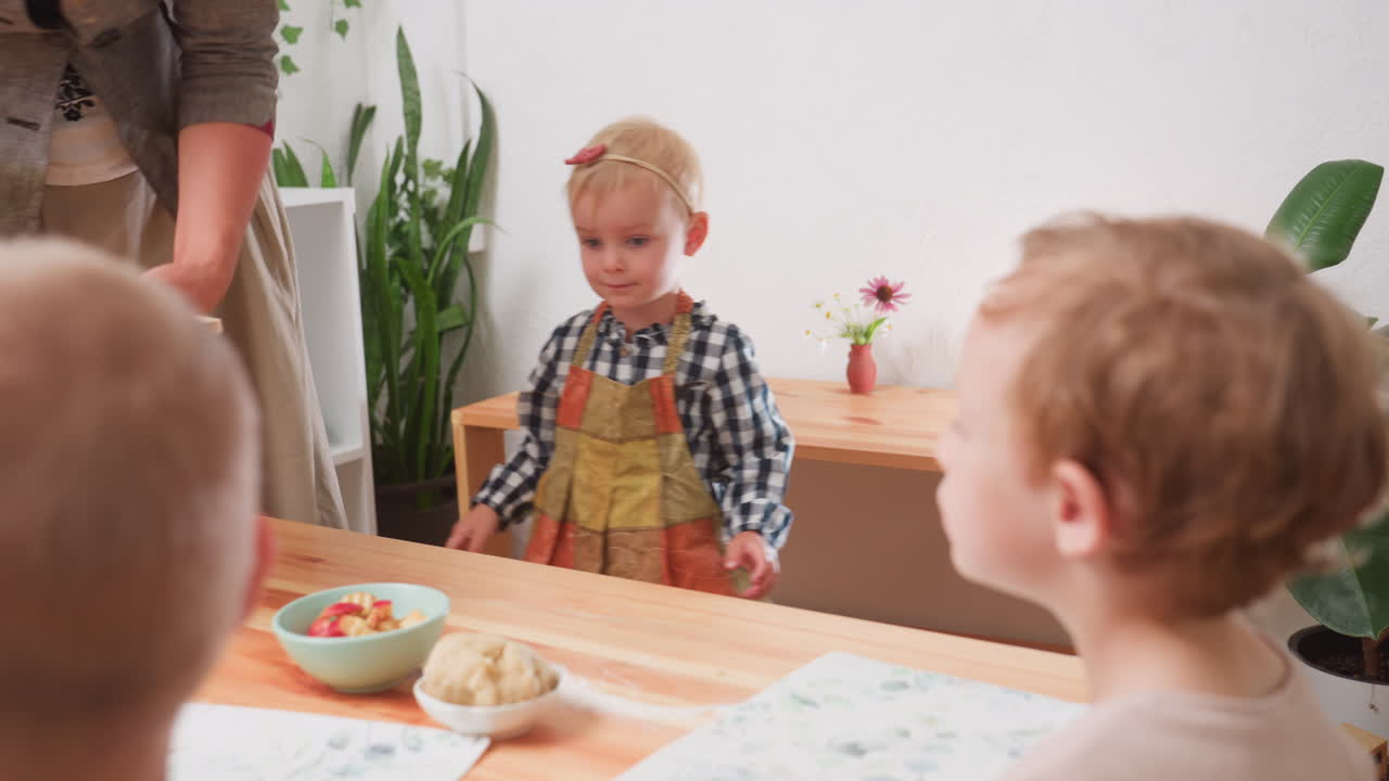 Bambini seduti attorno al tavolo che guardano l'insegnante riempire una ciotola mentre una bambina con un grembiule a scacchi porta un piatto di fettine di mela durante l'ora di lezione all'asilo