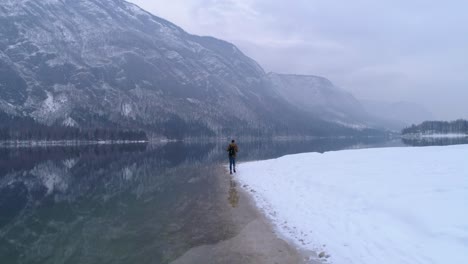 man walks alone, snowy bohinj lake reflecting mountains in distance, aerial flyover shot