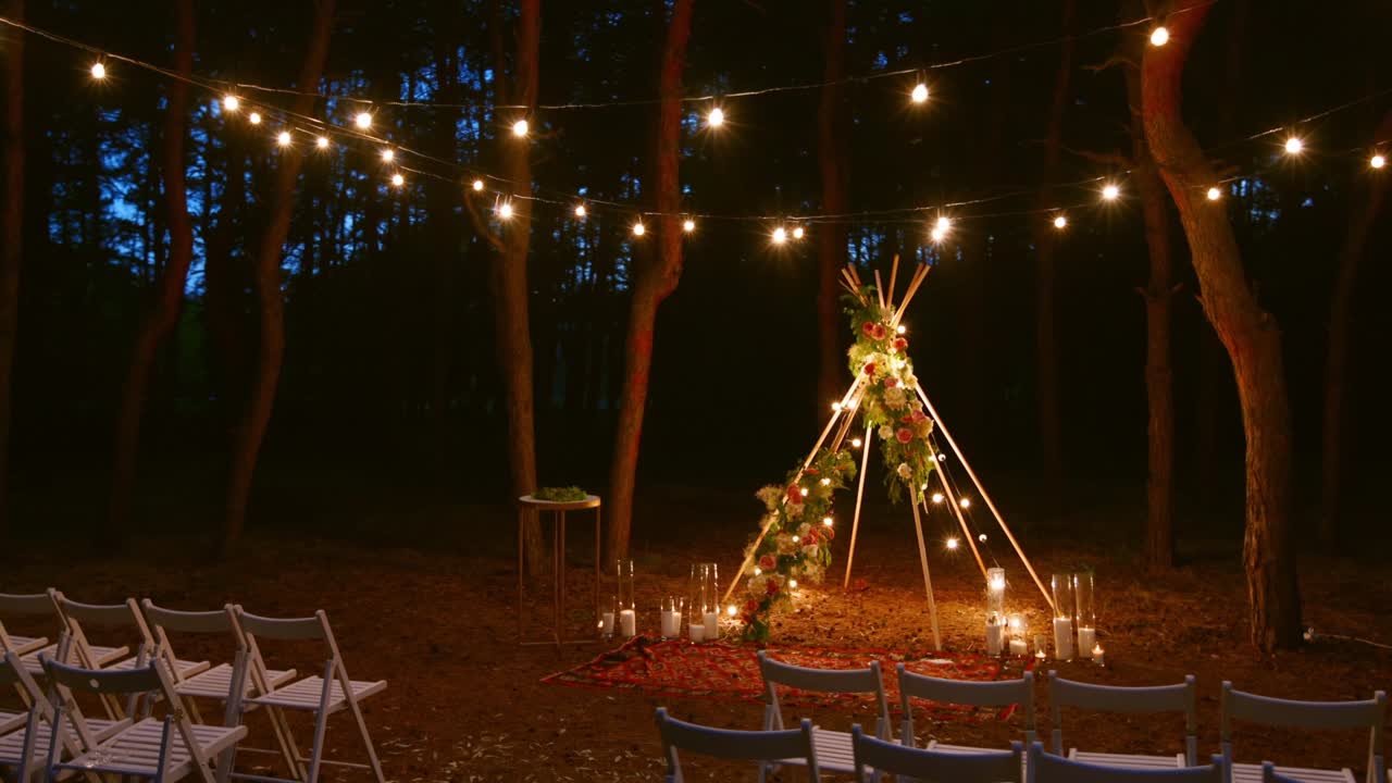 luces de cadena festivas iluminación en el arco de boho tipi decoración en el lugar de la ceremonia de boda al aire libre en el bosque de pinos por la noche. luces de cuerda vintage guirnaldas de bombillas que brillan sobre las sillas en la boda rural de verano